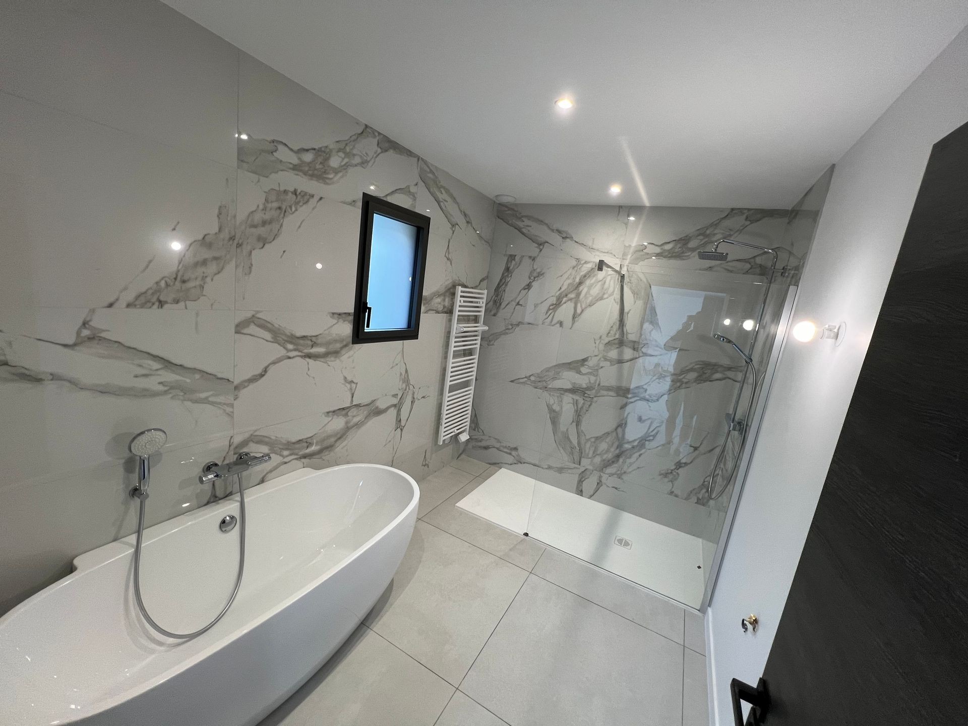 Modern bathroom with marble walls, oval bathtub, walk-in shower, and a small window.