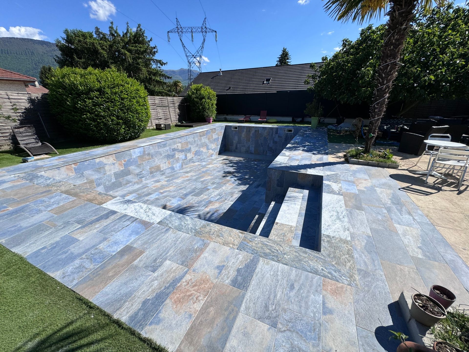 Empty tiled swimming pool in a backyard surrounded by plants and outdoor furniture on a sunny day.